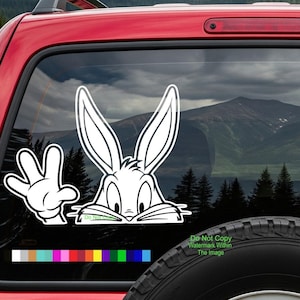 May include: A white cartoon rabbit sticker with a peace sign hand gesture. The rabbit has large ears and is peeking over a line. The sticker is on a red vehicle window with a mountain and forest background.