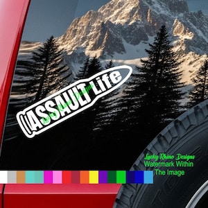 Op de afbeelding: Een wit en groene "ASSAULT Life" sticker wordt weergegeven op een rode voertuigruit. De sticker staat tegen een achtergrond van een bergketen en groenblijvende bomen. De band van het voertuig is zichtbaar in de rechter benedenhoek.