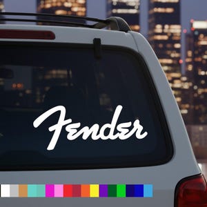 May include: White Fender decal on the rear window of a white car. The Fender logo is in a cursive font. The background features a blurred cityscape with illuminated buildings.