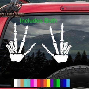 May include: White skeleton hand decals, each displaying the middle finger, are affixed to a vehicle's window. The text "Includes Both" is above the decals. The background shows a mountain range and trees.