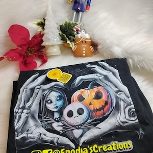 May include: Black t-shirt featuring a graphic of Jack Skellington, Sally, and a carved pumpkin. The design is framed by skeletal hands. Additional decorative items include a nutcracker, a gingerbread man, and a poinsettia.