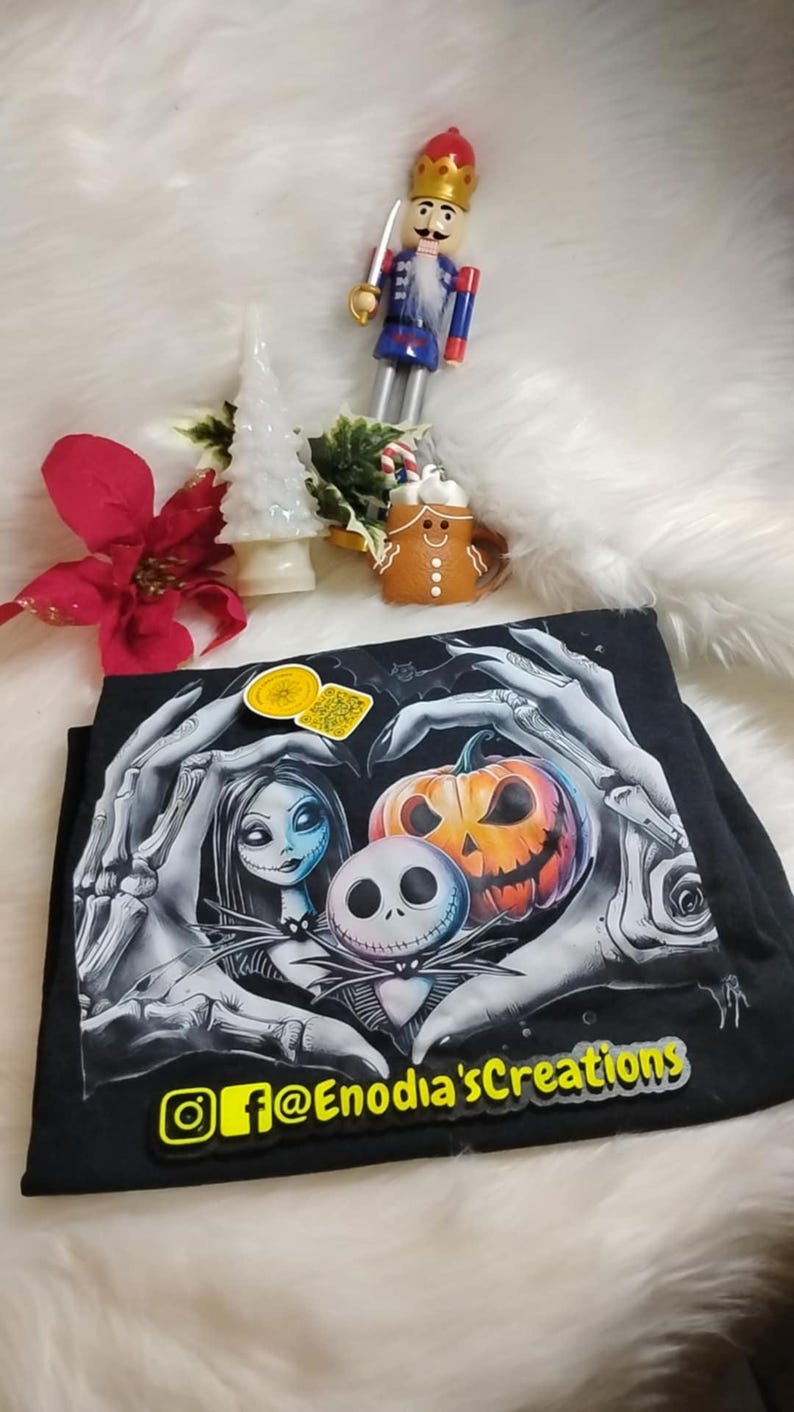 May include: Black t-shirt featuring a graphic design with characters from The Nightmare Before Christmas, including Jack Skellington, Sally, and a jack-o'-lantern. The shirt also has the text "@Enodia'sCreations". A wooden nutcracker and other holiday decorations are in the background.