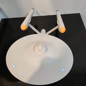 May include: A model of the U.S.S. Enterprise NCC-1701, a light gray spaceship with orange and white accents. The model features the ship's iconic saucer section and two warp nacelles. The text "U.S.S. Enterprise NCC-1701" is visible on the saucer.