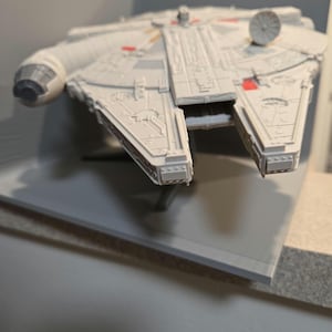 May include: A detailed model of the Millennium Falcon starship, predominantly light gray with red and yellow accents. The model features intricate paneling and a radar dish. The ship is displayed on a gray surface, showcasing its iconic design.