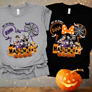 May include: Two gray and black t-shirts with a Mickey Mouse head design. The shirts feature a group of Disney characters dressed in Halloween costumes. The text on the shirts reads "Not so scary!" with a name, "John" and "Evva", and "Halloween".