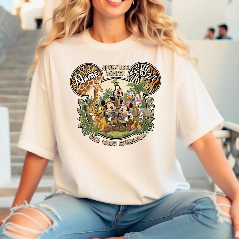 May include: White t-shirt featuring a colorful graphic of Disney characters, including Mickey and Minnie Mouse, in safari attire. The design includes the text "Name", "Adventure Awaits!", "2021", and "Wild Parks Expedition".