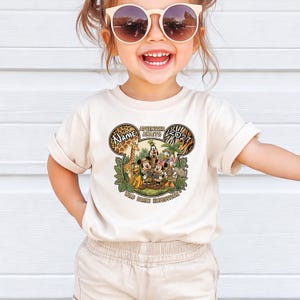 May include: A cream-colored t-shirt featuring a colorful graphic of Disney characters in a jungle setting. The design includes the text "Adventure Awaits" and "Wild Park Expedition". The shirt has short sleeves and is worn by a child.