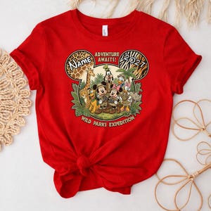 May include: A red t-shirt featuring a graphic of Disney characters in safari attire. The design includes the text "Adventure Awaits!" and "Wild Parks Expedition." The year "2021" is also displayed.