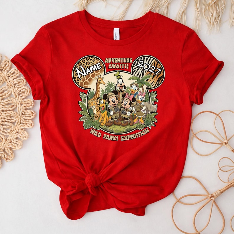 May include: A red t-shirt featuring a graphic of Disney characters in safari attire. The design includes the text "Adventure Awaits!" and "Wild Parks Expedition." The year "2021" is also displayed.