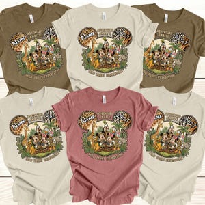 May include: A collection of t-shirts in various colors, including tan, brown, and rose. Each shirt features a graphic of Mickey Mouse and friends in safari attire, with the text "Adventure Awaits! 2027" and "Wild Parks Expedition."