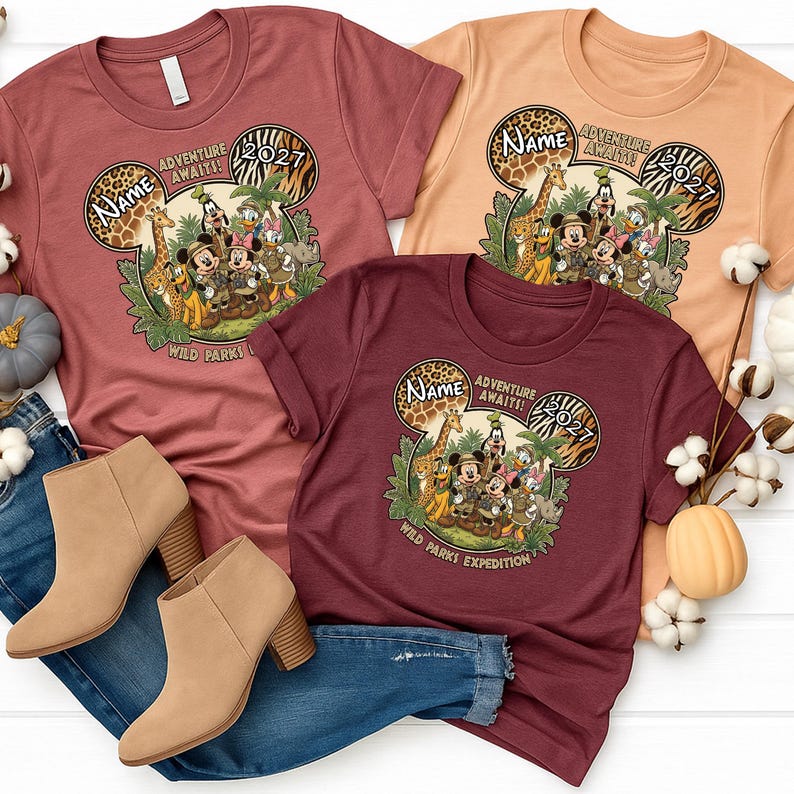 May include: Three t-shirts in shades of red and tan, featuring a Mickey Mouse-themed design. The design includes Mickey and friends, giraffes, and the text "Adventure Awaits! 2027" and "Wild Parks Expedition". Tan ankle boots and blue jeans are also in the image.