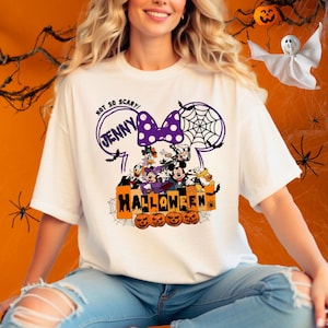 May include: White t-shirt with a purple and white Minnie Mouse ears design. The ears have a spiderweb and the text "Not So Scary! Jenny" inside. The design also includes Mickey Mouse, Donald Duck, Goofy, and Pluto characters with the text "Halloween" and pumpkins.