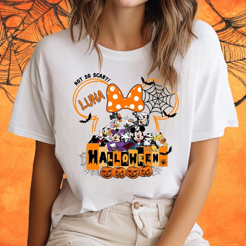 May include: White t-shirt with a Halloween design featuring Mickey Mouse and friends. The design includes a large Mickey Mouse ear with a bow, a spiderweb, and the text "Not So Scary! Luna Halloween".