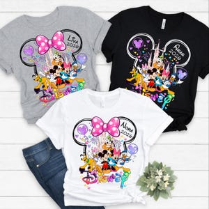 Op de afbeelding: Drie t-shirts met Disney-personageafbeeldingen. De shirts zijn grijs, zwart en wit. Elk shirt heeft een Mickey Mouse-hoofdomtrek met een roze stippen strik, de naam van een persoon en het jaar 2026. Het woord "Disney" is in kleurrijke letters geschreven.