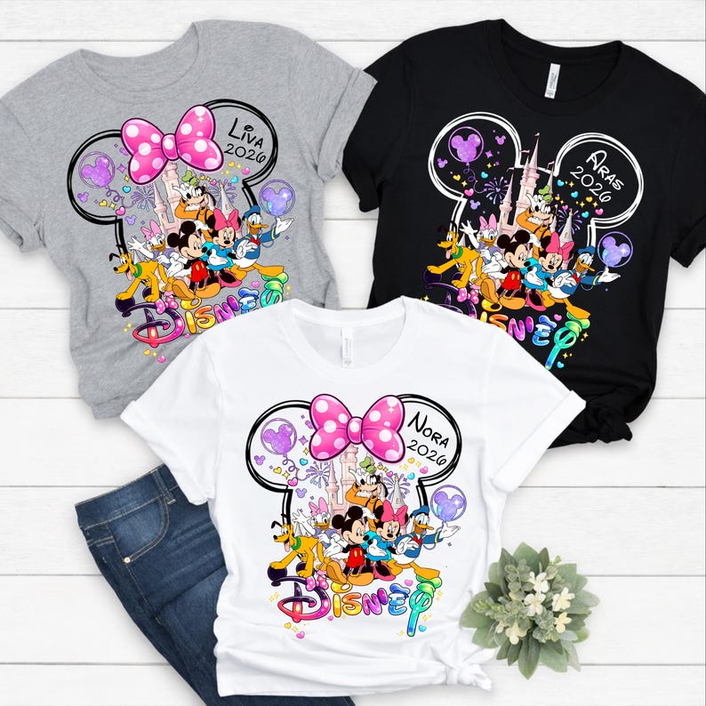 Op de afbeelding: Drie t-shirts met Disney-personageafbeeldingen. De shirts zijn grijs, zwart en wit. Elk shirt heeft een Mickey Mouse-hoofdomtrek met een roze stippen strik, de naam van een persoon en het jaar 2026. Het woord "Disney" is in kleurrijke letters geschreven.