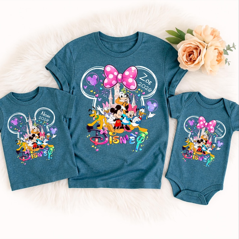 Op de afbeelding: Drie teal Disney-shirts. De grootste is een t-shirt met een roze strik, Disney-personages en de naam "Zoe 2026". De andere twee zijn een t-shirt en een romper, elk met een vergelijkbaar ontwerp en de namen "Noah 2026" en "Luna 2026".
