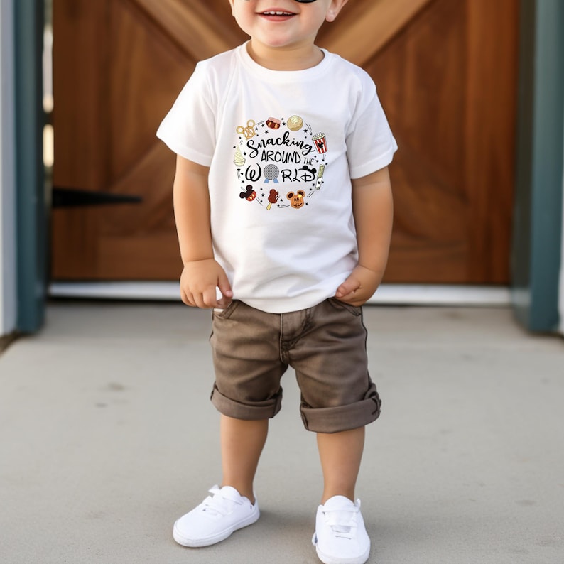 May include: A white t-shirt with a graphic of a Mickey Mouse head and the text "Snacking Around the World". The shirt is being worn by a young child wearing brown shorts and white sneakers.