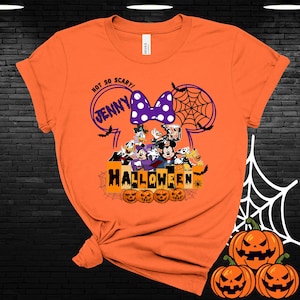 May include: Orange t-shirt with a Mickey Mouse Halloween design. The design features Mickey Mouse, Minnie Mouse, Donald Duck, Goofy, and Pluto. The text on the shirt says "Not So Scary! Jenny Halloween".