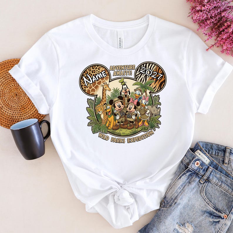 May include: White t-shirt featuring a colorful graphic of Disney characters in safari attire. The design includes Mickey and Minnie Mouse, Donald and Daisy Duck, Goofy, and other animal characters. Text includes "Name", "Adventure Awaits!", "2027", and "Wild Parks Expedition".