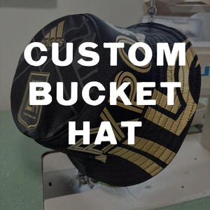 May include: Black and gold bucket hat with the text "CUSTOM BUCKET HAT" in white. The hat features a geometric pattern and a team logo. The hat is on a sewing machine.