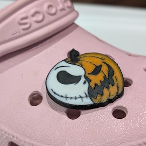 May include: A shoe charm featuring a split design of Jack Skellington and a jack-o'-lantern. The charm is black, white, and orange, and is attached to a light pink clog.