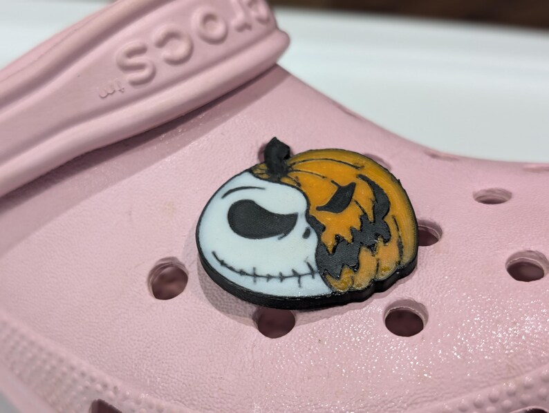 May include: A shoe charm featuring a split design of Jack Skellington and a jack-o'-lantern. The charm is black, white, and orange, and is attached to a light pink clog.