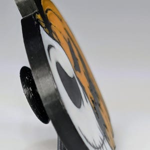 May include: A black, white, and orange circular decorative item featuring a skull design. The item is propped up on a black stand. The skull design is split in half with orange and black stripes on one side and a white skull on the other.