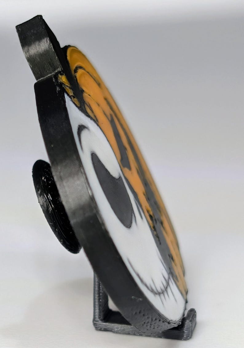 May include: A black, white, and orange circular decorative item featuring a skull design. The item is propped up on a black stand. The skull design is split in half with orange and black stripes on one side and a white skull on the other.