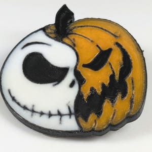 May include: A 3D-printed Halloween decoration featuring a split design. One half is a white skull with black eyes and a stitched grin, the other is an orange jack-o'-lantern with black cut-out features. The stem is black.