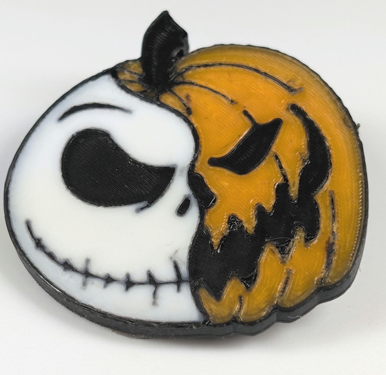 May include: A 3D-printed Halloween decoration featuring a split design. One half is a white skull with black eyes and a stitched grin, the other is an orange jack-o'-lantern with black cut-out features. The stem is black.