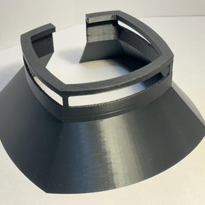 Op de afbeelding: Een zwart, 3D-geprint object in de vorm van een kraag. Het object heeft een brede, uitlopende basis en een verhoogd, geometrisch bovendeel met rechthoekige uitsparingen. Het ontwerp is hoekig en modern, geschikt voor cosplay of kostuums.