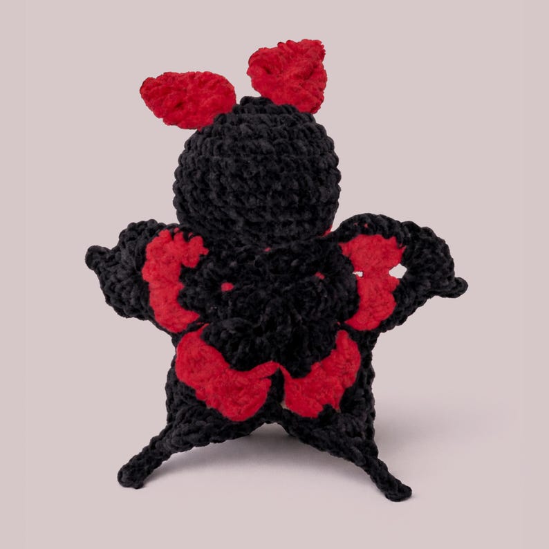 Puede incluir: Una mariposa hecha a mano con ganchillo, en negro y rojo. La mariposa tiene alas grandes con detalles rojos y dos antenas rojas. El cuerpo es negro y las alas est&aacute;n extendidas. El objeto artesanal se muestra sobre un fondo rosa claro.