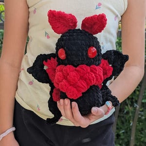 Puede incluir: Un peluche de polilla negra hecho a mano con detalles rojos. El peluche presenta ojos rojos, un pecho rojo y antenas rojas. La polilla se sostiene en las manos, mostrando la textura suave y la artesan&iacute;a detallada del juguete.