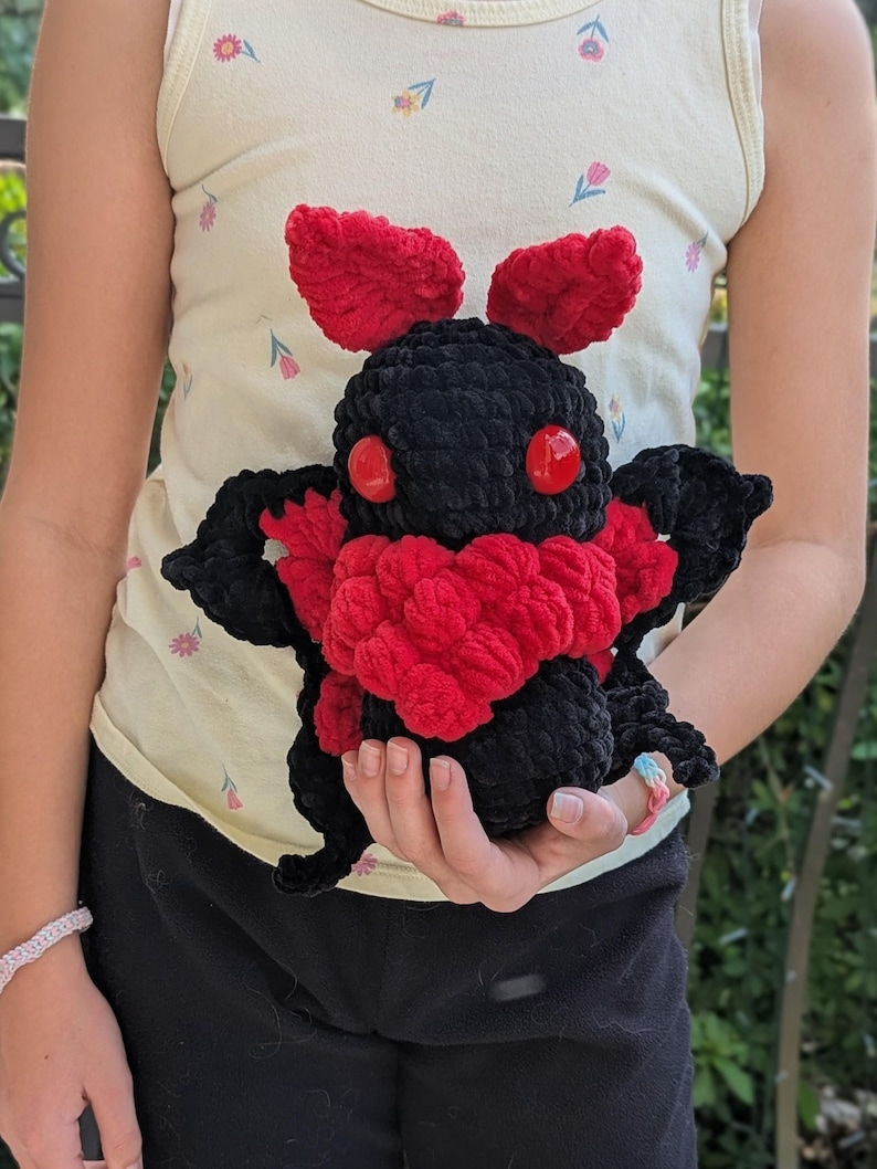 Puede incluir: Un peluche de polilla negra hecho a mano con detalles rojos. El peluche presenta ojos rojos, un pecho rojo y antenas rojas. La polilla se sostiene en las manos, mostrando la textura suave y la artesan&iacute;a detallada del juguete.