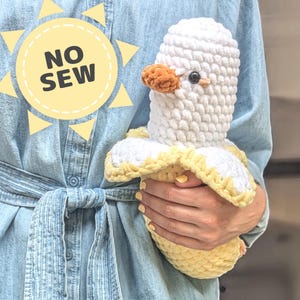 Könnte beinhalten: Ein gehäkeltes Bananen-Plüschtier mit weißem Körper und gelber Schale. Das Spielzeug wird von einer Person gehalten, die ein blaues Jeanshemd trägt. Der Text "NO SEW" ist auf einem gelben Sonnengrafik im Hintergrund sichtbar.