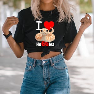 Op de afbeelding: Zwart T-shirt met de tekst "I ❤️ HoGGies" en een afbeelding van een slang met een rood hart. De slang is oranje en wit. Het shirt wordt gecombineerd met een blauwe jeans.