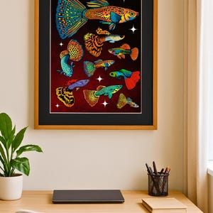 May include: A framed art print featuring colorful illustrations of various guppy fish species. The fish are depicted with vibrant hues of blue, orange, yellow, and green against a dark red background. The artwork is displayed on a wooden desk with a laptop and office supplies.