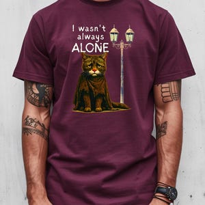 May include: Burgundy t-shirt featuring a dejected cat illustration with a red nose and the text "I wasn't always ALONE." The cat is positioned in front of a vintage street lamp. The shirt is a solid color.
