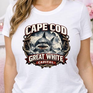 Pode incluir: Camiseta branca com um gráfico de um tubarão branco no oceano, com as palavras "Cape Cod Great White Capital" em um design circular. O design inclui âncoras e corda.