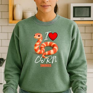 Könnte beinhalten: Grünes Sweatshirt mit dem Text "I ❤️ Corn Snakes" und einer Grafik einer rot-orangen Kornnatter. Das Design beinhaltet ein rotes Herz und das Wort "Corn" in einer stilisierten Schriftart.