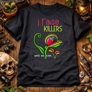 May include: Black t-shirt with a colorful graphic of a Venus flytrap and a fly. The text "i raise KILLERS" is above the plant, and "WATER. SUN. VICTIMS." is below. The design features a red, green, and yellow color scheme.