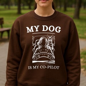 May include: A brown sweatshirt with the text "MY DOG IS MY CO-PILOT" in white. The graphic shows a dog in a car with a person driving. The person is wearing the sweatshirt and blue jeans.