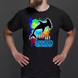 May include: Black t-shirt featuring a hammerhead shark silhouette with the text "Let's Get Hammered" in blue and white. The design includes colorful paint splatter accents in blue, green, yellow, and red.