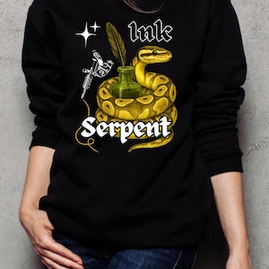 Może przedstawiać: Czarna bluza z grafiką przedstawiającą żółto-brązowego węża zwiniętego wokół zielonej butelki z tuszem. Napis "Ink Serpent" jest napisany białą czcionką. W projekcie uwzględniono również maszynkę do tatuażu i pióro.