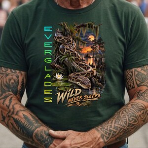 May include: A dark green t-shirt featuring a graphic of a large snake coiled on a tree branch in a swamp at sunset. The text EVERGLADES is printed vertically in light blue, and the text WILD NEVER SLEEPS is printed in yellow and white.