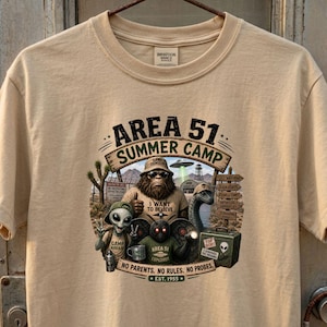 May include: A beige short-sleeve t-shirt featuring a graphic print titled Area 51 Summer Camp. The design shows a Bigfoot, an alien, and a moth-like creature with text including I Want To Believe, Area 51 Explorer, and No Parents. No Rules. No Probes.