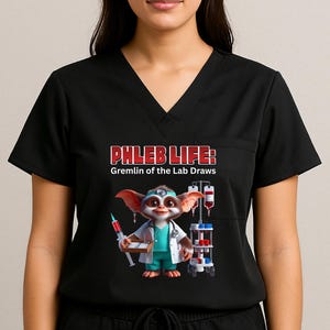 Puede incluir: Top de uniforme negro con cuello en V y bolsillo, con el texto "PHLEB LIFE: Gremlin of the Lab Draws". El gráfico muestra un gremlin con bata de laboratorio sosteniendo una jeringa y un dispositivo médico. La modelo lleva pantalones negros.