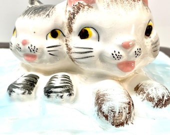 May include: A ceramic figurine featuring two white cats with brown and black markings. The cats have pink noses and ears, and yellow eyes. The figurine is on a light blue base with brown trim.