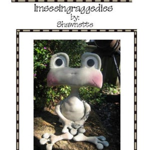 Puede incluir: Una caprichosa estatua de rana de hormigón gris llamada "Freddie Frog", de 35,5 cm de altura, con mejillas rosadas y ojos grandes y expresivos. La imagen incluye el texto "Imseeingraggedies by: Shawnette."