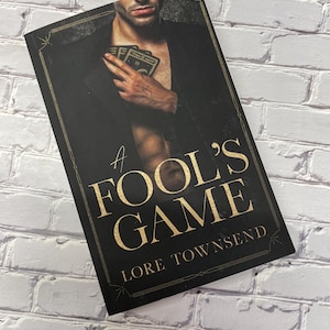 Puede incluir: Una cubierta de libro negra con letras doradas que dicen "A Fool's Game" y "Lore Townsend". La cubierta presenta a un hombre sosteniendo cartas de juego en su pecho.